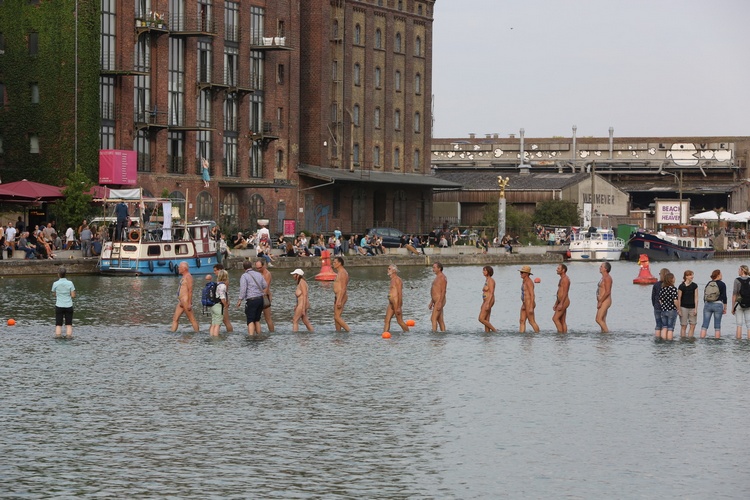 Elf blote performers lopen langzaam over de waadplaats naar het midden van de havenkom